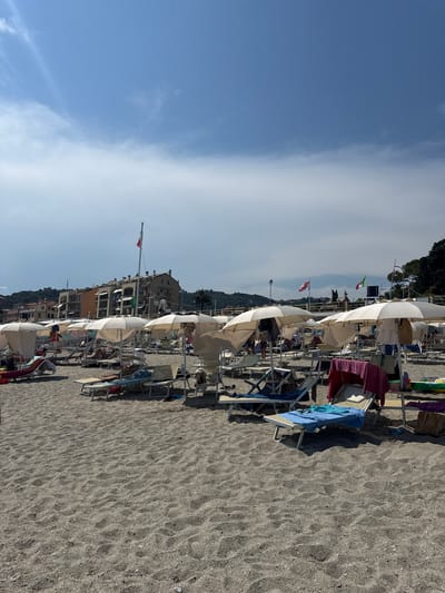 The image depicts a crowded beach scene in Finale Ligure, Italy. Numerous white beach umbrellas are set up on a sandy shore, with many lounge chairs beneath them. Some chairs are occupied by people relaxing, while others are draped with towels and clothing. In the background, buildings with a Mediterranean architectural style are visible, along with lush green hillsides. Several flags are flying from poles, including what appears to be the Italian flag. The sky is partly cloudy with bright sunlight. The foreground is dominated by the textured sand of the beach.