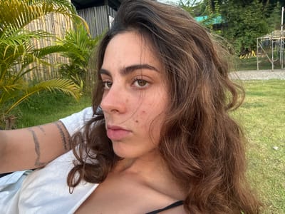 The image depicts a close-up, slightly low-angle view of a single adult female in an outdoor setting within Ban Mae Ping, Thailand. The subject has medium-brown, wavy hair and brown eyes, with several small moles or freckles visible on her face, particularly on her right cheek below the eye and on her chin. Her lips are slightly parted, and her expression is neutral. She is wearing a white, loose-fitting top, under which a black strap is partially visible on her left side. A tattoo consisting of multiple horizontal bands and script is present on her left forearm. The background immediately behind her consists of green grass. To her left, tropical palm-like foliage with green and yellow fronds is visible, behind which a light-colored, possibly bamboo or wooden, slatted structure is present. To her right, a gravel or dirt path is visible beyond the grass, with dense green trees and foliage further in the distance. A metallic, scaffold-like structure is also visible on the far right in the background. The lighting suggests daylight conditions.