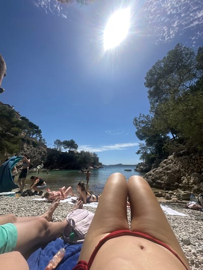 The image is taken at a beach in Peguera, Spain, featuring a sunny sky above. The scene shows a rocky coastline with trees and buildings in the background, framing a cove with clear, turquoise water. In the foreground, a group of people is relaxing on the pebbled beach. A person wearing a red swimsuit is prominently positioned, while other individuals are sunbathing or standing near the water's edge. Beach towels and personal belongings are scattered around.