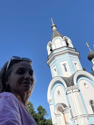 In St. Ozerki, Russia, on a clear day with a bright blue sky, a woman with blonde hair is captured in a low-angle selfie. She is wearing white sunglasses perched on her head and a light purple t-shirt, with a hoop earring visible in her left ear. The background features a white and light blue church with onion domes and golden crosses. The church's architecture includes arched windows and decorative detailing. Trees are visible behind the woman and to the left of the church.