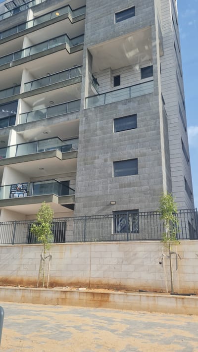 The image depicts the exterior of a modern, multi-story building under a clear blue sky, seen from a low-angle perspective looking upwards. The building features a mix of architectural styles and materials.

The upper section of the building has a light-colored, possibly stucco or stone, facade with horizontal textured lines and several small, dark-recessed windows. Below this, a large section of the building is clad in dark gray, rectangular stone or tile blocks, arranged in vertical rows. This section features several prominent, square, dark-paned windows, some appearing to have blinds or shutters.

Further down, the building incorporates numerous balconies with clear glass railings, creating a staggered or stepped appearance. The underside of these balconies and the walls behind them are a light, neutral color. Some balconies show reflections of the sky and surroundings. On one of the lower balconies, a white "For Sale" sign is prominently displayed, largely visible in the cropped images.

Adjacent to the building, a dark metal fence or railing runs parallel to its base, with small green trees or bushes planted along it. Beyond the fence, to the right, there is a paved area made of light-colored, possibly sandy-toned, rectangular stones arranged in a pattern, with some darker patches. A lower wall made of lighter, square blocks with visible rust-colored staining is also present behind the fence.

The scene is brightly lit by natural sunlight, indicating daytime with good weather. There are no people visible, and no discernible activity is taking place.

Visible text on a white "For Sale" sign on one of the balconies (as seen in the cropped images) reads in Hebrew: "למכירה" (For Sale), "דופלקס 6 חדרים" (6-room duplex), "רחוב [unclear street name] קריית גת" ([street name] Kiryat Gat), confirming the location as Kiryat Gat. It also lists a phone number: "050-8451660", and additional descriptions such as "דופלקס מפואר" (Luxurious duplex), "חדש" (New), and "כניסה מיידית" (Immediate entry).