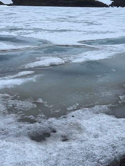 This media file captures an aerial or overhead view of a partially frozen body of water in Poschiavo, Switzerland, dominated by various formations of ice and snow.

The scene depicts a winter landscape characterized by a mix of dark, open water, smooth ice, and textured snow-covered ice. On the left side of the frame, a dark bank, possibly rocky or muddy, with some irregular white patches that could be snow or ice, borders the water. The main body of the image showcases a dynamic surface where clear, dark-grey water or thin ice coexists with thicker, opaque white ice and snow.

The ice presents diverse textures: some areas are relatively smooth and reflective, while others are rough, granular, and heavily ridged, suggesting layers of accumulated frozen material. Particularly on the right side, the surface is densely covered in a thick, frothy, or bubbly-textured snow and ice, creating a rugged topography.

Visible objects include a small, dark, round piece of debris, possibly a stone, resting on the textured snow on the middle-right. Another very small, light-colored, rectangular item, perhaps a piece of plastic or paper, is embedded in the ice near the bottom-left.

There is no discernible human activity or event taking place; the image is a still capture of a natural environment shaped by cold weather conditions. The lighting is diffused, suggesting an overcast day, and imparts a cool, muted palette to the scene. No visible text is present.