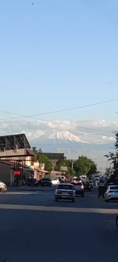 The image shows a street scene in Ayntap, Armenia with Mount Ararat visible in the background. The foreground consists of a street with moving cars. A building with a visible number "216" is located on the left side of the street. Power lines are visible in the sky.