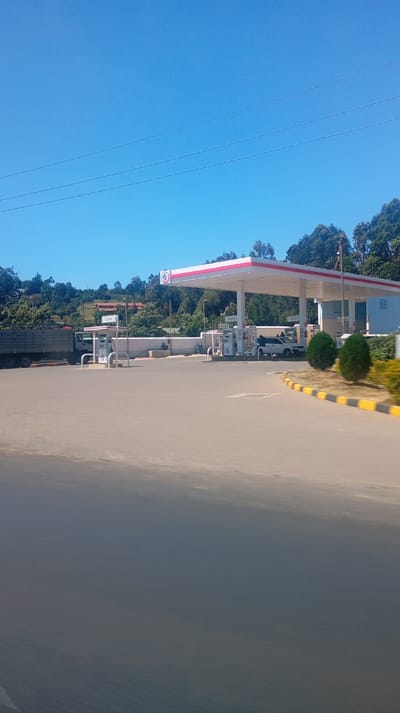 A daytime view features a TotalEnergies gas station with multiple pumps under a white and red canopy. A white pickup truck is parked near the pumps. A truck loaded with a dark material is adjacent to the station. Background elements include lush green hills with scattered houses and a clear, blue sky with visible power lines. A road occupies the foreground. Yellow and black curb markers define an edge of the station.