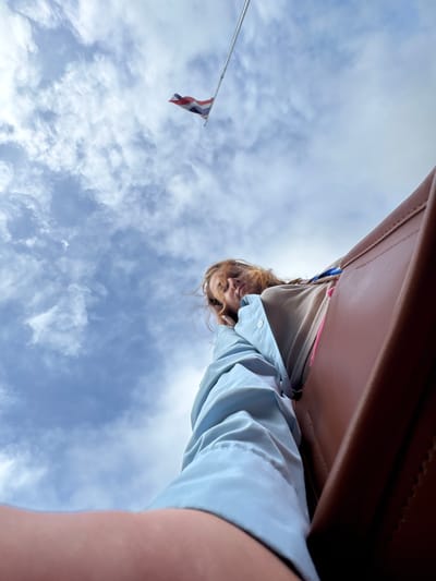 The image, taken from a low-angle perspective looking upwards, shows a young woman with reddish-blonde hair, wearing a light blue collared shirt over a tan sweater. She is leaning back and looking towards the sky, with her hand resting near her face. To her right, a portion of a brown leather surface, possibly a seat or luggage, is visible. In the upper portion of the image, a flag with horizontal red, white, and blue stripes, identified as the flag of Thailand, flies against a cloudy blue sky. The specific location in Tambon Wang Prachan, Thailand, is not discernible from the visual information provided.