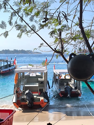 This image captures a tranquil scene at a beach in an unspecified location in Indonesia, likely a tropical island. In the foreground, two small motorboats are docked on a sandy shore, their hulls partially submerged in the clear turquoise water. One boat, closer to the viewer, is white with an orange trim and features an outboard motor at the stern. The other boat, slightly further back, shares a similar design. Both vessels are adorned with Indonesian flags, adding a touch of local color.

Behind the boats, the ocean stretches out, dotted with distant islands and smaller boats. The water is a vibrant blue, with gentle waves lapping at the shore. Lush green foliage, including leafy branches and some seed pods, hangs from trees in the foreground, framing the view and casting dappled shadows. A large, dark, spherical object, possibly a buoy or mooring weight, hangs from a branch in the right foreground. The sky is a clear, bright blue, indicating a sunny day. The overall impression is one of a peaceful, idyllic coastal setting.
