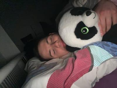A person is lying in bed next to a stuffed panda toy. The person's eyes are closed, and their face is resting against the panda. The bed has patterned bedding in shades of gray, blue, and pink. The image appears to be taken in a dimly lit room. There is no information in the image about the location being in Benningen, Germany.