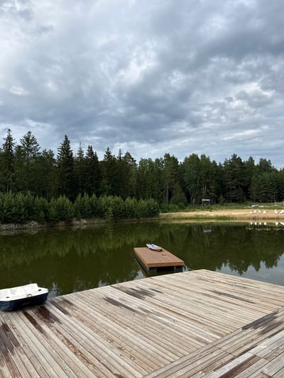 The image depicts a serene lake scene with a focus on a wooden dock and a surrounding natural landscape. The dock, constructed from weathered wooden planks, occupies the right side of the frame and extends into the water. A small, dark-colored object, possibly a bag or a case, rests on the dock near its edge. A white boat is moored to the dock, partially submerged in the calm, dark water.

The lake itself is a prominent feature, its surface reflecting the dense green forest that lines the opposite shore. The trees are a mix of tall pines and other deciduous species, creating a lush and verdant backdrop. A narrow strip of sandy beach is visible between the water's edge and the treeline. Beyond the beach, more trees and some white structures, possibly beach chairs or changing booths, are faintly visible.

The sky above is overcast with a layer of textured gray clouds, suggesting a temperate day, possibly late afternoon or early morning given the soft, diffused light. The overall mood is tranquil and peaceful, emphasizing the natural beauty of the setting. There is no visible text within the image. The location is identified as Sosnovka, Russia.