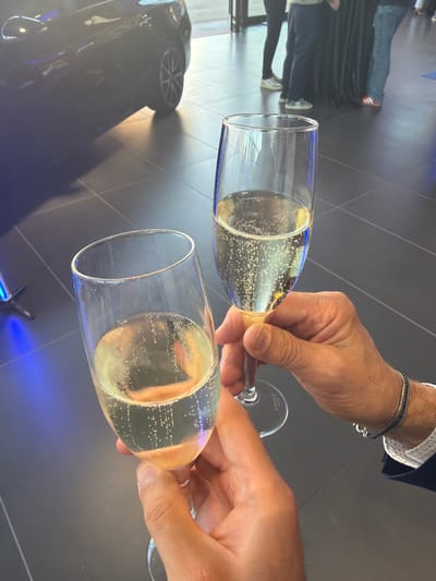 Two champagne flutes filled with a bubbly, pale yellow liquid are being held by two people. The flutes are clear glass with thin stems. The background is dark and appears to be a tiled floor. A dark-colored car is visible in the upper left corner of the image. In the background, there are several people standing, some wearing dark clothing and others in casual attire. The scene suggests a celebration or event, possibly at a car dealership or a formal gathering. The image does not provide any specific visual cues to indicate a location in Parma, Italy.