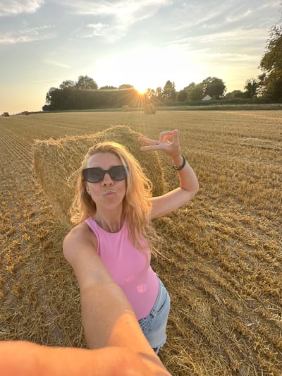 A woman is taking a selfie in a harvested wheat field, with bales of hay in the background. She is wearing a pink tank top and denim shorts, and has blonde hair and sunglasses. She is making a peace sign with her right hand and puckering her lips. The sun is setting, casting a warm glow over the scene. The location is the city of Trouville-Alliquerville, France.