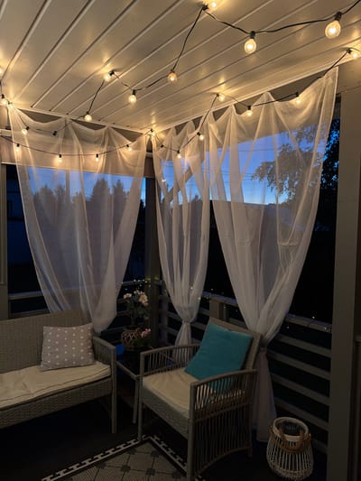 The image depicts a covered outdoor patio or porch area, likely at dusk or night, as evidenced by the warm glow of string lights and the darkening sky visible through sheer white curtains. The patio is furnished with two woven wicker chairs and a matching sofa. The sofa has a light-colored cushion and a grey throw pillow with white star patterns. The chair in the foreground has a light-colored seat cushion and a turquoise accent pillow. A dark rug with a geometric pattern is visible on the floor. A small, dark-colored table sits between the furniture, holding a potted plant with light-colored flowers and another smaller pink flower. A white wicker lantern with rope handles is placed on the floor next to the chair. The ceiling of the patio is paneled with white wood, and the string lights are attached to it, casting a warm illumination. The sheer curtains are draped from the ceiling, creating a soft, ethereal effect and obscuring the view of the exterior, though hints of trees and a building are visible through them. The railing of the patio is dark-colored and appears to have another strand of small fairy lights along it. The overall impression is one of a cozy and inviting outdoor relaxation space, potentially located in the city of Alyoshkino DP, Russia, given the context provided. There are no people visible in the image.