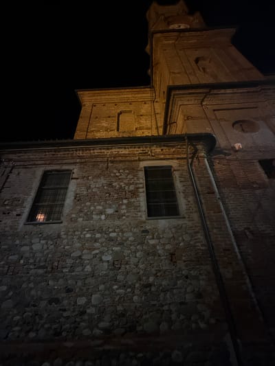 The image shows a low-angle, nighttime view of a brick building, likely a church or bell tower, in Lagnasco, Italy. The building is constructed from reddish-brown bricks and features prominent architectural elements such as cornices and window frames. Two rectangular windows with grates are visible on the lower section of the building, with light reflecting within one of them. A large downspout runs vertically along the side of the building. The upper portion of the image reveals a section of a taller structure, possibly a bell tower, with a clock face partially visible. The sky is dark and devoid of any celestial bodies. The overall scene is dimly lit by artificial light, casting shadows and highlighting the textured surfaces of the brickwork.
