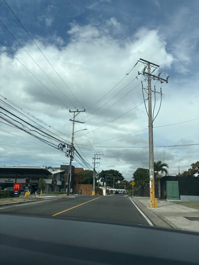 The image shows a view from a car looking down a paved road in San José, Costa Rica at the intersection of Avenida 10 and Calle 51. The road is empty, and there are power lines running overhead. The power lines are supported by several utility poles. The intersection is marked with yellow lines on the road and a yellow diamond-shaped sign on the left side. There are buildings and trees on either side of the road. The buildings are mostly single-story and have a commercial appearance. The sky is overcast with white clouds. The sun is shining. The image is taken from the perspective of a person sitting in a car.