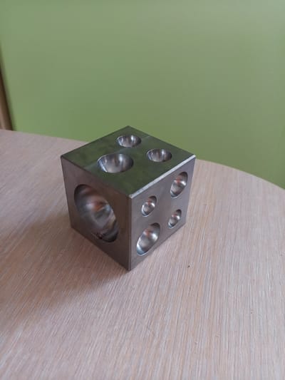 The image shows a metallic cube with several hemispherical indentations on its faces. The cube is positioned on a light-colored, wood-grained surface, possibly a table. The background is a solid, light green wall. The lighting appears to be from the front and slightly from the right, casting subtle shadows on the surface and within the indentations of the cube. The object itself appears to be a tool or a component for crafting or manufacturing. There are no people or animals visible in the image. The image is a still photograph, and there is no discernible action taking place. The scene is indoor. There is no information in the image to indicate its location in Vilyuysk, Russia.