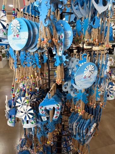 The image shows a close-up, eye-level, vertical shot of a retail display of blue and white souvenirs. The souvenirs are primarily made of painted wood or metal and are suspended from a black metal rack by rough twine. Many of the items are shaped like windmills, horseshoes, seahorses, and anchors, often adorned with floral patterns. Some also feature beads in various colors, including blue, white, yellow, orange, and pink, as well as "evil eye" charms. The background is blurred but suggests an indoor retail environment with shelves and other merchandise visible. The location is identified as Acıpayam, Turkey, suggesting this is a shop in that city. No people are visible in the frame.
