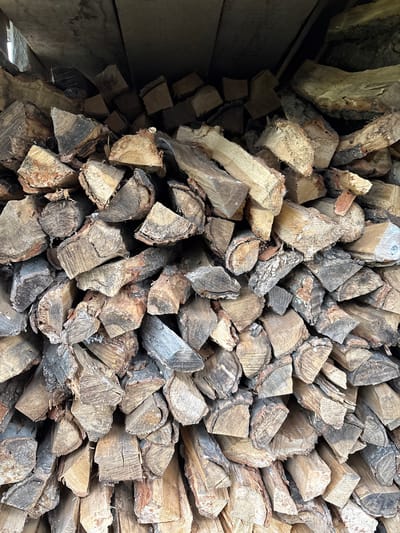 The image is a close-up, vertical shot of a large stack of cut firewood. The wood is primarily composed of logs split into various sizes, with some larger, rougher pieces visible. The bark is still attached to many of the pieces, and the wood shows evidence of age and weathering, with variations in color from light brown to dark gray. The grain of the wood is clearly visible, with rings and cracks indicating the material's organic nature. The firewood is stacked densely, filling the frame, with the top of the stack meeting a dark, flat surface, possibly the underside of a roof or shelter. The lighting is somewhat dim, casting shadows and highlighting the textures of the wood. There are no people or animals present in the image. The scene appears to be an outdoor storage area for firewood. The location is specified as Siniy Gay, Russia.