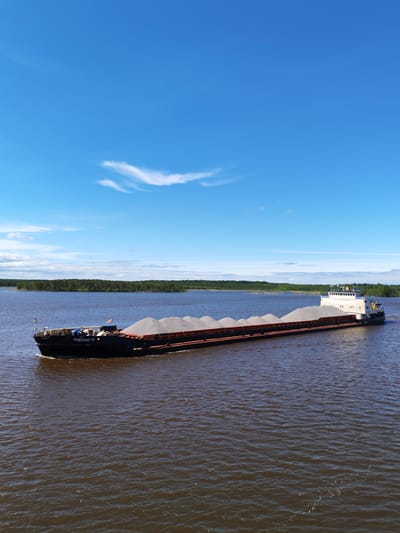 The image shows a large barge traveling on a wide, calm river. The barge is filled with a cargo of gray granular material, likely gravel or sand. It is moving from left to right in the frame. On the left side of the barge, the name "НЕВСКИЙ 18" is visible in white lettering. The rear of the barge features a white superstructure with a yellow railing.

The river water is dark, and the sky above is a bright blue with a few wispy clouds. In the background, a strip of green forest lines the horizon, suggesting the river passes through a natural area.

Based on the name "НЕВСКИЙ 18" and visual characteristics, the barge is likely traveling on a waterway in Russia. Given the prompt to include the location Novodevich'e, it can be concluded that the photo was taken in Russia, likely near Novodevich'e. The river could potentially be part of the Volga or another major Russian waterway system.