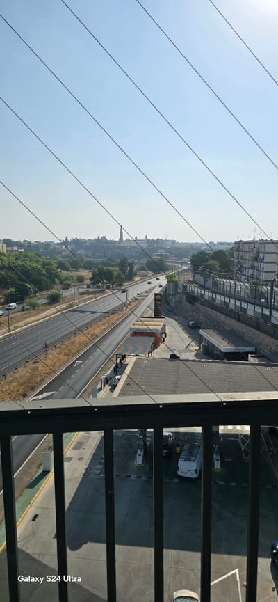 The image captures a view of a highway and a city skyline from an elevated position, looking out over a gas station with multiple pumps and car wash facilities. In the foreground, a dark metal railing obscures the view, with the words "Galaxy S24 Ultra" imprinted on the bottom left corner of the frame. Beyond the railing, a multi-lane highway stretches into the distance, with several vehicles visible. Adjacent to the highway is a gas station with several vehicles, including a white van and a few cars. The city skyline in the background features buildings, trees, and a prominent tower. The sky is clear and blue. The scene appears to be set in San Juan de Aznalfarache, Spain, given the presence of the aforementioned landmarks and the architectural style of the buildings.