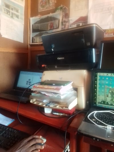 The image shows a desk in Auchi, Nigeria (7.063, 6.274) with a printer, two laptops, and a stack of papers. The printer is a Canon, and the laptops are both open and have their screens visible. The laptops are both connected to the printer with cables. A person's right hand is visible in the lower right corner of the image. They are using a black computer mouse. The desk is brown and wooden. The wall behind the desk is brown and covered in corkboard. There are a few papers and drawings pinned to the wall. The scene is dimly lit. The overall appearance suggests a business or office environment.