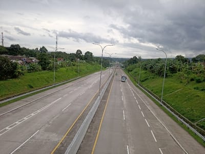 The image depicts a multi-lane divided highway in Caringin, Indonesia, viewed from an elevated perspective, likely an overpass, under an overcast sky. The highway consists of at least three lanes in each direction, separated by a concrete median barrier with yellow safety lines. The road surface is light grey concrete with white dashed and solid lane markings. Modern streetlights with curved arms are uniformly spaced along both sides of the highway.
In the right-hand lanes, a white and green passenger bus travels away from the viewer, followed by a smaller green vehicle further in the distance. Other indistinct vehicles are visible in the far distance. Guardrails line the outer edges of the highway.
The immediate environment features grassy embankments rising from the road. Beyond these, dense tropical vegetation, including numerous banana plants and various trees, covers rolling terrain. In the upper left background, several residential structures with light-colored walls and dark roofs are partially visible amidst the foliage. A tall, lattice-style communication tower stands on a distant hill to the far left. The horizon is characterized by hills covered in dense greenery. No identifiable people are visible.