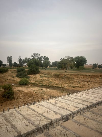 A landscape view dominated by a hazy sky. The terrain features a mix of vegetation and exposed earth. Trees and shrubs dot the mid-ground, hinting at a natural or rural setting. In the foreground, a concrete structure with visible rebar suggests construction or engineering work, contrasting with the natural backdrop. The image appears to be taken from an elevated perspective, potentially from atop the concrete structure itself.