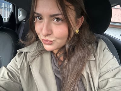 A young adult female is centered, posing for a selfie from the front passenger seat of a vehicle. She has long, wavy, dark brown hair, brown eyes, and a puckered lip expression with light, glossy lipstick. A small silver hoop piercing is present in her right nostril, and a similar hoop is in her left helix. A circular gold-toned dangle earring with a patterned design adorns her right earlobe. She wears a light olive-green trench coat over a gray, textured long-sleeved garment. The vehicle's interior shows black synthetic or leather upholstery. Raindrops are visible on the exterior side window. The out-of-focus background reveals a light-colored building facade with dark horizontal lines and a metal railing structure. This scene is located in the city of Nesso, Italy.