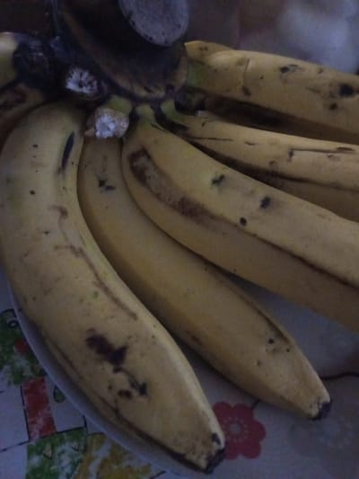 This image, taken in Nam Mat, Laos, displays a close-up view of a cluster of approximately six to seven ripe bananas. The bananas are yellow with varying degrees of brown and black spots, streaks, and blemishes indicative of ripening and some surface imperfections. Their stems, fused at the top, are dark brown to black. One central banana shows a lighter, fibrous interior where its stem may have been partially detached. The bananas rest on a patterned surface, likely a tablecloth, featuring abstract blocks of green, white, yellow, and red, with a distinct pinkish-red floral design visible in the lower right corner. No people, specific actions, or interactions are present in the scene, which appears to be an indoor setting under ambient or artificial lighting.