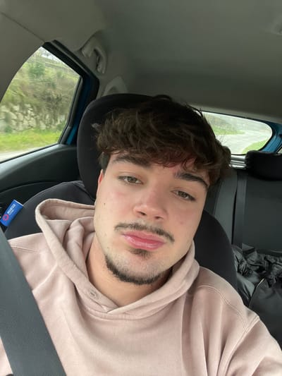 The image captures a male individual in a close-up selfie taken inside a vehicle within Lamego, Portugal. The subject has brown, wavy hair, light brown eyes, a styled mustache, and a goatee. He is wearing a light pink/beige hooded sweatshirt and is secured by a dark seatbelt across his left shoulder and chest. Slight redness is visible on his forehead, and his lips are slightly pouted as he looks directly at the camera.

The vehicle's interior features dark seating, a grey headliner, and blue-colored window frames. A white object marked with a "Sparco Corse" logo is attached to the seatbelt pillar on the left. A dark, crumpled item, possibly a garment, rests on the seat behind the subject to his right.

Through the vehicle's windows, a natural outdoor environment, consistent with a rural or suburban area in Lamego, Portugal, is visible. The left window shows a green and brown hillside with a partial stone wall. The rear window reveals a paved surface, likely a road, receding into further vegetation.