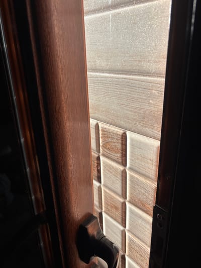 The scene depicts an interior perspective looking through a partially opened door in the city of Razhdak, Bulgaria. On the left, a dark brown, wood-grained doorframe is prominent, with a black, metallic door handle visible on the door itself, which is slightly ajar. Beyond the opening, a wall covered in textured ceramic tiles is brightly illuminated by direct natural light, highlighting suspended dust particles in the upper portion of the light beam. The upper tiles feature a light beige, horizontally striped wood-grain pattern. Below this, the tiles transition to a three-dimensional pattern of alternating light cream and brown rectangular blocks with rounded edges, creating a visually textured surface. The right edge of the door opening reveals the dark metal frame and strike plate of the doorjamb. No individuals or active interactions are present in the image.