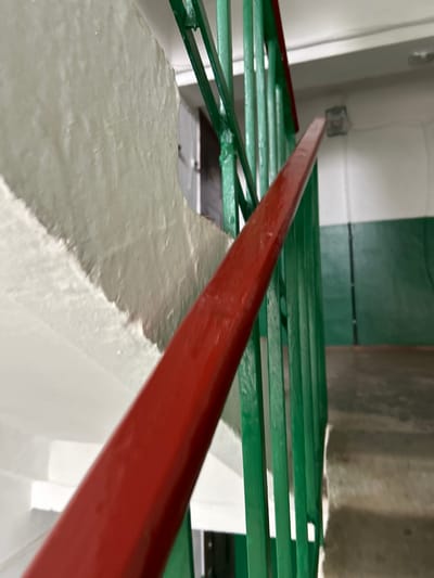 The image displays an interior stairwell within a building, reportedly located in the city of Chernomurovskii, Russia. The perspective is an oblique downward shot, primarily focusing on a section of the staircase railing and the adjacent wall.

The central subject is a metal stair railing. Its top handrail is painted a glossy, vibrant red. Beneath the red handrail, a series of vertical, rectangular metal balusters are painted dark green. These balusters descend towards the right side of the frame, indicating the downward direction of the staircase.

To the left of the railing, a textured concrete wall runs parallel to the stairs. This wall exhibits a two-tone paint scheme: the upper section is off-white, characterized by visible peeling paint and an uneven surface texture. The lower section, partially obscured by the railing, is painted a solid dark green.

On the far right, concrete steps with visible risers and treads descend, also painted in a dark green hue on their vertical surfaces. In the background, an additional section of the wall is visible with the same white upper and green lower paint scheme. A rectangular, flush-mounted light fixture is affixed to the upper wall/ceiling junction in the background. A doorway with a partially visible white door is present behind the railing on the left side of the frame. No individuals or active interactions are depicted. The overall environment suggests an older, utilitarian building interior with visible signs of wear and aging paintwork.