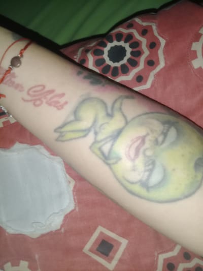 A close-up shot focuses on a person's forearm, which is adorned with two tattoos. The first tattoo is a cartoonish depiction of a figure resembling a pear or an apple, with human-like features such as eyes, eyelashes, a nose, and lips, all shaded in a greenish-yellow hue. The second tattoo, located closer to the wrist, appears to be a red-inked inscription. The forearm is also encircled by a thin red bracelet with a brown bead. The arm rests on a patterned fabric, featuring red, black, and white geometric shapes. The background includes a green surface and another piece of patterned fabric. The location is not discernible from the visual information.