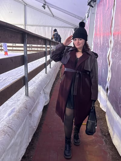 A woman in a black beanie and a dark, leather-accented trench coat stands inside a white tent. She wears dark stockings and black lace-up boots. A black purse hangs from her shoulder. Behind her, a metal structure with snowy coverings stretches into the distance. The tent's roof is translucent, allowing light to filter through. Another person is visible in the background, near the tent's entrance. The ground is a muted red color.