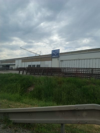 A long industrial building with a corrugated metal roof and white siding stands under a cloudy sky. A crane is visible on the left side of the building. A blue sign with the logo "ARMABAU" is mounted on the facade. In front of the building, piles of rebar are stacked. Lush green grass and weeds grow in front of the rebar piles and extend to a gray metal guardrail in the foreground. The location is the ARMA-BAU company in Kaplice, Czechia.