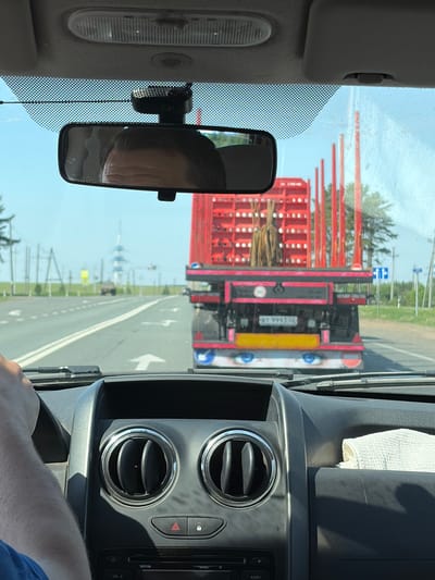 A view from inside a vehicle, possibly a car or truck, showing a red logging truck directly ahead on a road in what appears to be daylight conditions. The logging truck is carrying logs, and its rear license plate is partially visible, reading "BT 9993 02". A speed limit sign with "90" is also visible on the truck. The road has white painted directional arrows. In the rearview mirror, a reflection of a person's face can be seen. The interior of the vehicle is visible, including the dashboard, air vents, and a white cloth or towel resting on the dashboard. In the distance, a large structure resembling a ship is seen on the horizon, suggesting a coastal area. There is a place called "Ильинское, Russia" that could be the location where the photo was taken. The logging truck is travelling away from a ship.