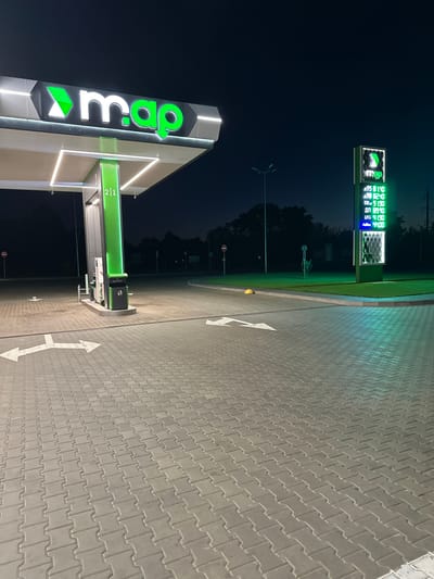 The image shows a gas station at night in Syrnyky, Ukraine. A large "map" logo is illuminated on the left side of the frame, above a covered gas pump area. A digital price board displaying fuel costs is visible in the background on the right. The ground is paved with gray interlocking bricks. The sky is dark, and a few trees are silhouetted against it. No people are visible in the scene.