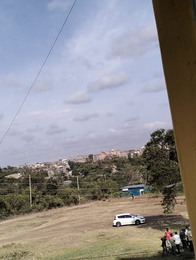 A daytime outdoor scene captures a town's edge with various buildings nestled among trees under a partly cloudy sky. An open dirt area in the foreground features a white car parked nearby. A group of people stand on the grass. Utility poles stretch across the scene. A section of a yellow building is visible on the right side of the frame, partially obscuring the view.