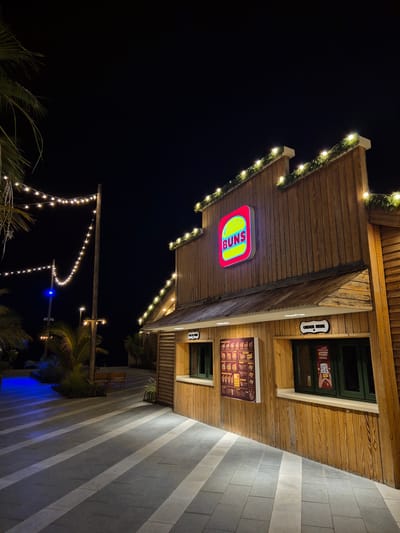 The image depicts a wooden-clad food establishment at night, likely in Fujairah, United Arab Emirates, given the context. The building has a rustic, cabin-like appearance with a sloped roof and wooden plank siding. A prominent red and yellow neon sign, shaped like a hamburger with the word "BUNS" inside, is mounted on the upper facade. String lights with white bulbs are draped along the roofline and a tall wooden pole to the left, casting a warm glow.
The establishment features two service windows. The left window displays a menu board with various food items categorized. The right window, with dark green trim, shows a QR code and text, possibly for ordering or information. A sign above this window reads "ORDER HERE" in a stylized font.
The ground in front of the building is paved with large, grey, rectangular tiles, arranged in a diagonal pattern, with lighter grey stripes running through them. To the left, illuminated by a vibrant blue light, is a walkway or seating area with palm trees and more string lights in the distance. Two wooden benches are visible in the mid-ground, suggesting an outdoor seating space. The overall atmosphere is festive and inviting, illuminated by artificial lights against a dark night sky. No people are visible in the frame.