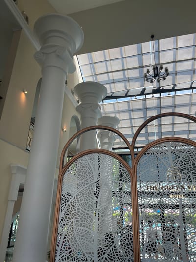 The image captures the interior of a building, likely a hotel lobby, featuring an atrium with a large glass roof. Prominently visible are several tall, white Corinthian columns that extend upwards towards the ceiling. In the foreground, a decorative screen with an intricate white pattern, possibly representing branches or foliage, and a bronze frame divides the space. Through the screen, the words "THE GREEN VIEW HOTEL" are discernible, suggesting the location. A multi-armed chandelier hangs from the ceiling. The overall ambiance suggests a well-lit, spacious interior with classical architectural elements. The image does not show any people. The location is identified as Bang Sao Thong Subdistrict, Thailand.