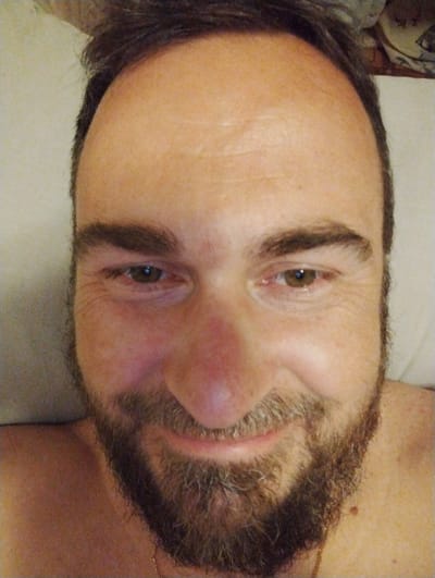 The image is a close-up selfie of a man with brown hair and a beard. He is looking directly at the camera, and his face is well-lit. The background appears to be a light-colored surface, possibly a bed or couch. There are some wrinkles visible on his forehead and around his eyes. The man is wearing a thin gold chain around his neck. No specific location in the city of Ширяево (ильинский), Russia can be identified from the image.