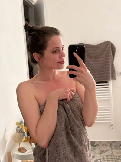 A woman is taking a selfie in what appears to be a bathroom. She is holding a black smartphone in her right hand, and the phone is positioned to capture her face and upper body. She is wrapped in a grey striped towel. Her hair is pulled back. The background shows a white wall, a grey towel hanging on a rack, a white radiator, and a patterned tile wall. In the bottom right corner of the image, there is a child's highchair. The scene does not appear to be in Perugia, Italy.