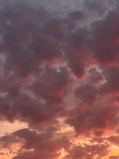 The image depicts a dramatic sky filled with turbulent clouds, exhibiting a gradient of colors from deep purples and grays at the top to fiery oranges and pinks towards the bottom. The clouds are dense and textured, with some areas appearing illuminated by the setting sun. There are no visible people, subjects, objects, or actions. The environment is solely the sky. The location in Rotmanka, Poland, is not visually discernible from the image itself; this information is external to the visual content.