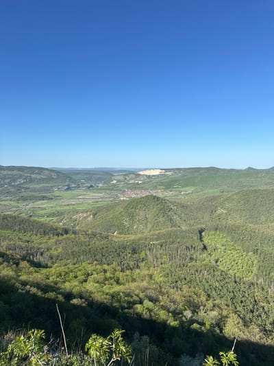 The image depicts a landscape view from a high vantage point in or near Bilka, Bulgaria. The sky is clear and blue, occupying a significant portion of the left side of the frame. Rolling hills and mountains covered with dense green vegetation dominate the right side of the image, extending into the distance. In the valley below, a small village or town is visible, with agricultural fields and patches of open land interspersed among the hills. Some trees are close to the edge of the frame. No people are visible.