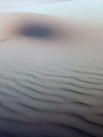 A close-up shot depicts a blurred landscape, reminiscent of rolling sand dunes or textured fabric. Dark, wavy lines traverse the lower portion, suggesting ridges or folds. A prominent, diffused dark spot sits atop a smoother, lighter area, possibly a shadow or variation in texture. The overall tone is muted and indistinct, emphasizing subtle gradations of light and shadow.