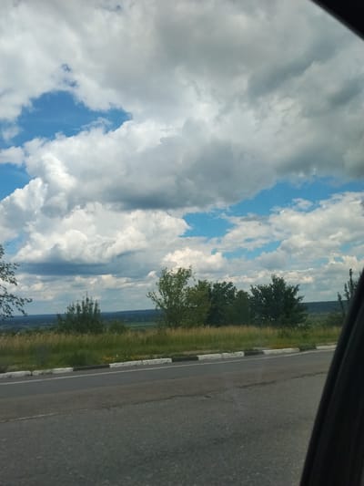 The image shows a view from inside a vehicle looking out at a landscape. In the foreground, the edge of the car's interior is visible on the right side of the frame. A paved road runs horizontally across the lower portion of the image, with a concrete curb separating it from a grassy area. Beyond the road and grass, there are trees and a distant view of a valley or rolling hills under a cloudy sky. The sky is a mix of blue patches and large, white and grey clouds. The scene appears to be a rural or semi-rural setting, possibly on the outskirts of a town or city.