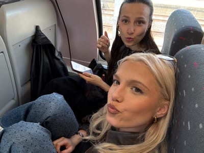 Two women are taking a selfie on what appears to be a train. The woman in the foreground has blond hair, is wearing hoop earrings, a brown leather jacket and a bracelet. The woman in the background has dark hair and is wearing large drop earrings. They are seated on a train with grey seats. The background shows a view of train tracks. In the background, a black leather jacket is hanging on a hook.
It's not possible to determine the exact location within Vilvoorde, Belgium, based on the image alone. More information would be required to pinpoint the specific train line or area.
