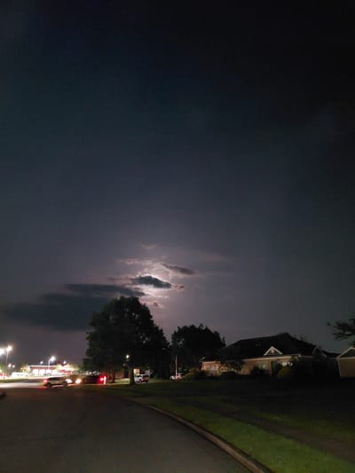 This is a nighttime photograph taken in Nicholasville, Kentucky, United States of America. The image captures a street scene with a dramatic sky. The sky is dark, with a visible indication of lightning behind a band of clouds, illuminating them from behind.
In the foreground, a paved road curves from the lower left to the lower right of the frame. The road appears to be residential or a quiet street. A neatly maintained green lawn runs along the right side of the road.
On the right side of the image, a low-lying residential building with a dark roof is visible. The building has several windows, some of which appear to be lit from within. The architecture suggests a modern suburban home. To the right of this house, a part of another building, possibly a garage or extension, is visible.
On the left side of the image, there are streetlights and the facade of a commercial building, possibly a gas station or convenience store, with illuminated signage. Several vehicles are present on the road, including cars with their headlights and taillights on, suggesting they are either driving or parked. One car in the middle of the frame has its brake lights illuminated.
A large, mature tree with dense foliage stands prominently in the middle ground, obscuring part of the residential building on the right. Another tree is visible in the far left, behind the commercial building. The overall atmosphere of the image is atmospheric, due to the stormy sky and the nighttime lighting.