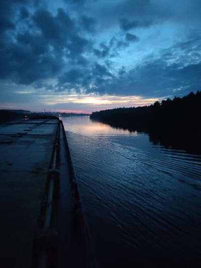 The image depicts a twilight scene over a wide river, possibly during dawn or dusk. Dark, ominous clouds dominate the upper portion of the sky, with streaks of pale orange and pink near the horizon, suggesting the last light of day or the first light of dawn. The water's surface is calm, reflecting the muted colors of the sky and creating gentle ripples from movement.
On the left side of the frame, a long, flat deck, likely part of a barge or a stationary structure, occupies a significant portion of the foreground. The deck appears to be made of concrete or a similar material and shows signs of wear. Several metal fixtures and railings are visible along the edge of the deck.
The right side of the image features a densely wooded shoreline, silhouetted against the sky. The trees are dark and indistinct due to the low light conditions. In the distance, along the river, several navigational lights are visible, indicated by small, colored points of light—red and green—emerging from the darkness. These lights suggest the presence of buoys or other markers in the waterway.
There are no people visible in the image. The overall atmosphere is serene, yet with a touch of somberness due to the dark clouds and the twilight. The location is identified as Bessonovo, Russia, implying this is a scene on a river in or near that region.