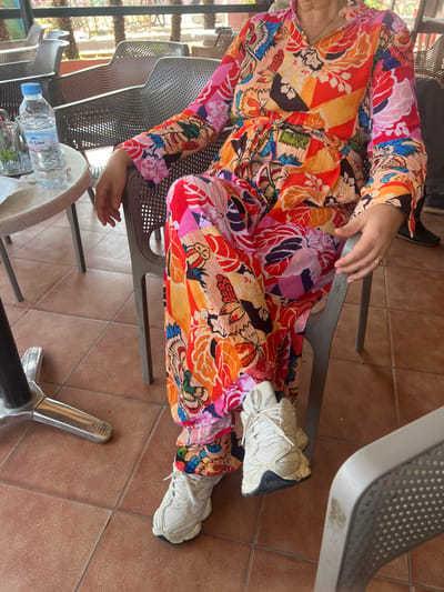 An individual is seated at an outdoor cafe in Mechra Bel Ksiri, Morocco. The person, viewed from the upper torso to the feet, wears a vibrant, multi-colored two-piece garment featuring a floral and abstract pattern in shades of orange, red, pink, blue, green, and black. The long-sleeved top has ruffled cuffs and a V-neck, and the trousers are loose-fitting. White athletic sneakers with chunky soles are worn; the right foot is slightly elevated and resting near the left. A thin gold-colored ring is present on the left ring finger. The individual is seated in a light grey, perforated plastic chair. To the left, a round light-colored table with a silver metal pedestal base holds a plastic bottle of 'Sidi Ali' mineral water, a clear glass containing a greenish liquid, and a white napkin. The flooring consists of reddish-brown square tiles. In the blurred background, additional similar cafe chairs and green foliage indicate an outdoor patio or terrace setting under daylight conditions.