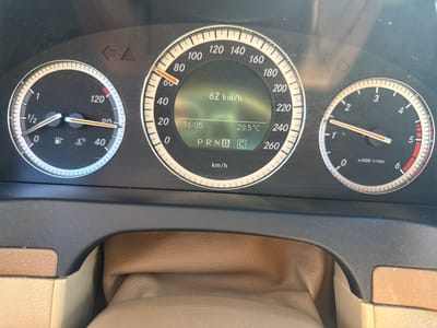 The image displays the dashboard of a car, likely a Mercedes-Benz, with its instrument cluster prominently featured. The dashboard is dark, with a textured surface. The central display shows a speed of "62 km/h", the time "16:05", and a temperature of "28.5°C". Below this, the gear selector is shown as "P R N D C". To the left of the central display is a fuel gauge and a temperature gauge for the engine. To the right is a tachometer, indicating "x1000 1/min". There are also illuminated indicator lights above the instrument cluster. The car's interior appears to be upholstered in tan leather. The image is taken from the driver's perspective. No information is available about the specific location within Sotirya, Bulgaria.