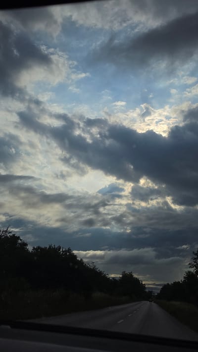 The image depicts a roadside view from a vehicle's perspective, traveling on a paved road towards a distant horizon. The road is bordered by dense, dark foliage, primarily trees and vegetation, on both sides. The sky above is dramatic, filled with a mix of heavy, dark clouds and patches of lighter, brighter sky where sunlight filters through. The overall lighting suggests it is either early morning or late afternoon, possibly with an impending storm. There are no visible people or significant human activity beyond the implication of travel on the road. The location is specified as being in the vicinity of Liliak, Bulgaria, which places the scene within a natural, possibly rural or semi-rural, Bulgarian landscape. The atmosphere is moody and overcast, with the dark clouds dominating the upper portion of the frame.